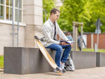 Junger Mann sitzt mit Skateboard auf Bank und notiert in Notizbuch