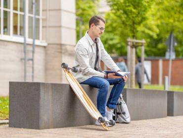 Junger Mann sitzt mit Skateboard auf Bank und liest in Notizbuch