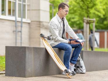 Junger Mann sitzt auf Bank mit Skateboard und notiert in Notizbuch