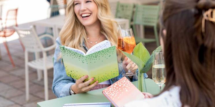 Frau lacht mit Notizbuch und Cocktail in Hand