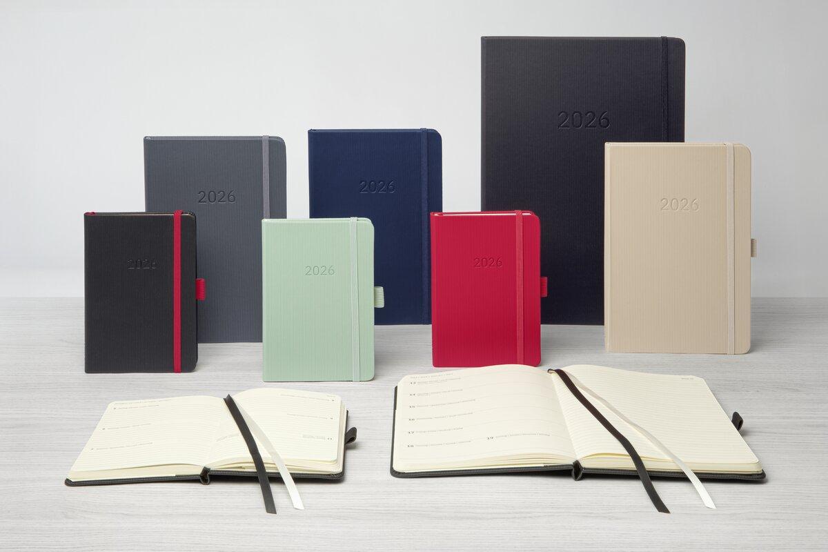 Verschiedene farbige Kalender 2026 stehen auf einem Holztisch Zwei geöffnete Kalender liegen davor