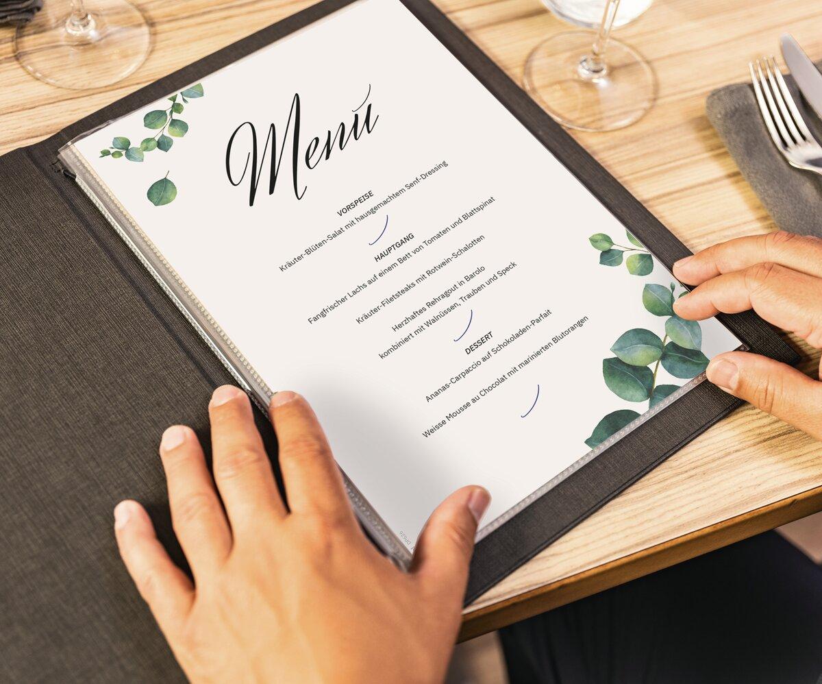 Menu in a black cover with eucalyptus design on wooden table, held by hands