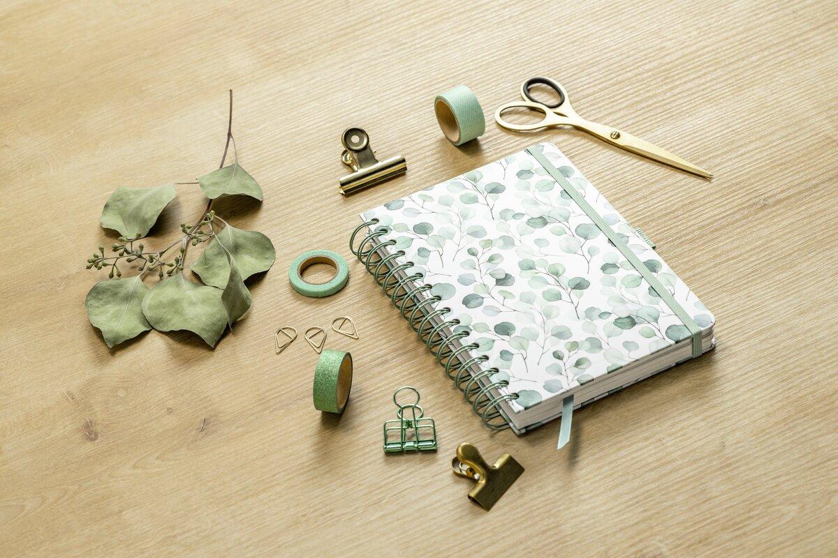 Notebook with plant pattern surrounded by paper clips, adhesive tapes, scissors, dried leaves and binder clip on wooden surface