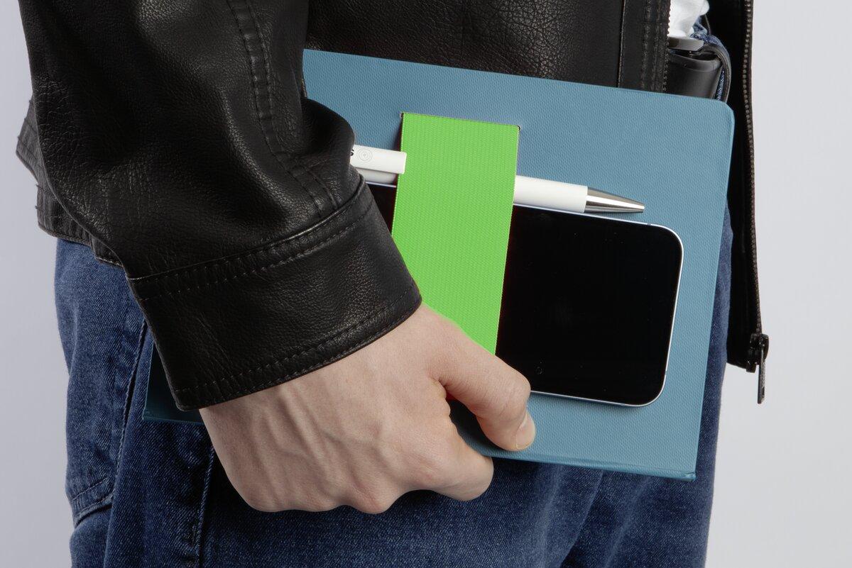 Person hält ein blaues Notizbuch mit grünem Band, Smartphone und Stift