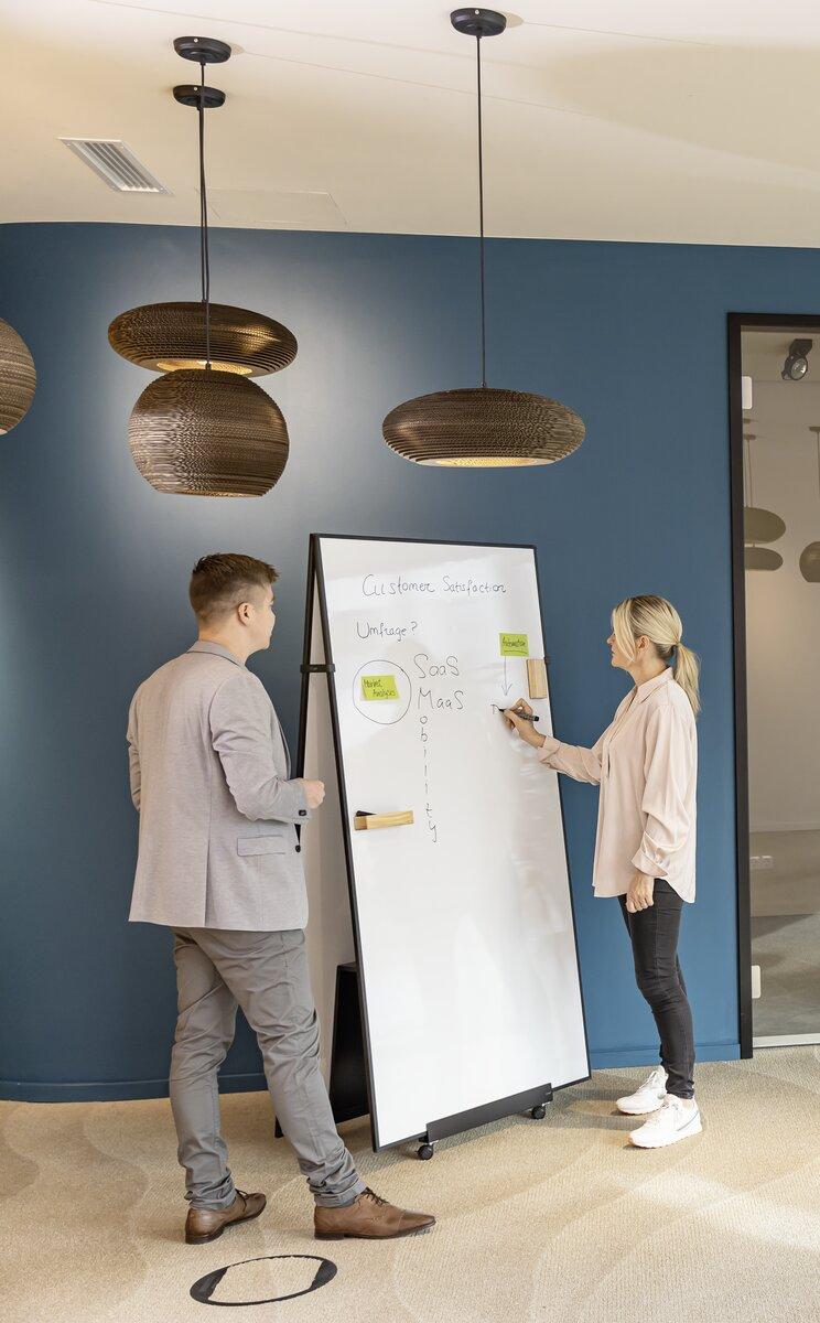 People interacting on a whiteboard in an office ceiling lamps and blue wall in the background