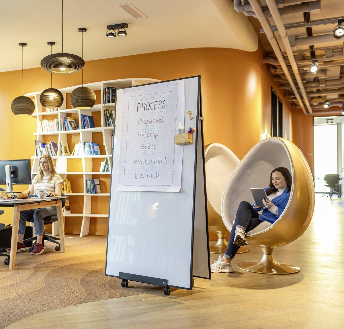 Open office with standing board and shelf, two people: one at the desk, one in an armchair