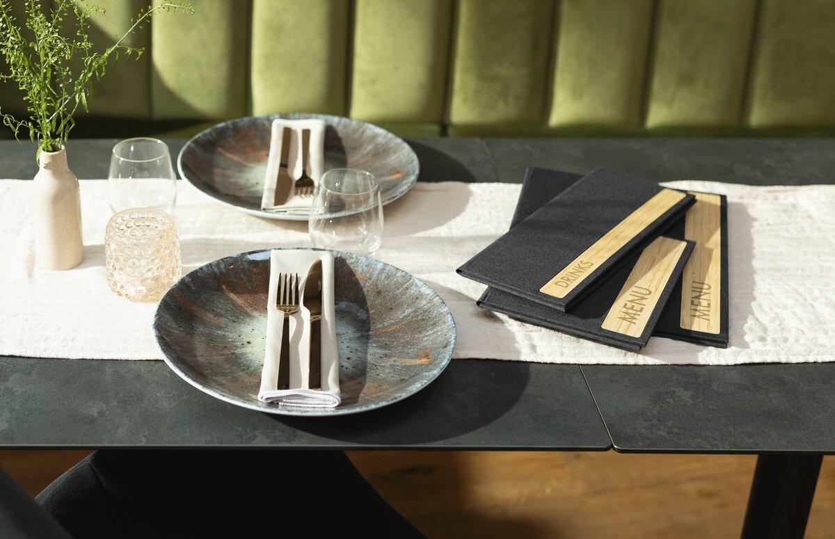 Table set with plates, cutlery, glasses and menu cards Vase with plant and upholstered green bench in the background