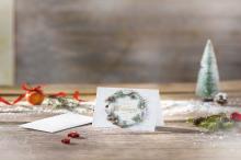 Christmas card with wreath motif, next to it envelope In the background an orange ball with a red ribbon and a small snow tree