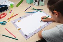 Child painting with green pen on white sheet surrounded by colorful writing material