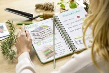 Notizbuch mit Skizzen eines Hochbeets und Pflanzplan Person blättert von Hand, neben Blumengabel und Rosmarin