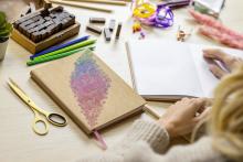 Notizbuch, Buntstifte und Hände beim Öffnen eines leeren Buches auf Holzuntergrund