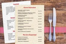 Speisekarte mit Getränken und Schmankerln, daneben Messer und Gabel auf Holztisch