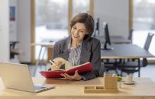 Frau sitzt lächelnd am Schreibtisch, hält ein aufgeschlagenes Notizbuch, Laptop und Tasse daneben