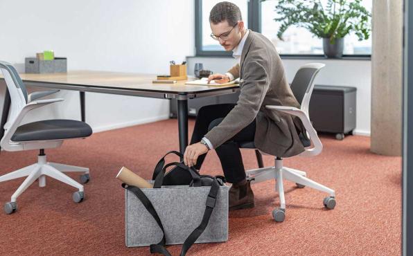 Mann sitzt an Schreibtisch und holt Arbeitsutensilie aus grauer Desk Sharing Bag auf dem Boden